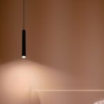 Schwarze Röhrenpendelleuchte, die einen warmen Lichtkegel auf eine braune Wand wirft