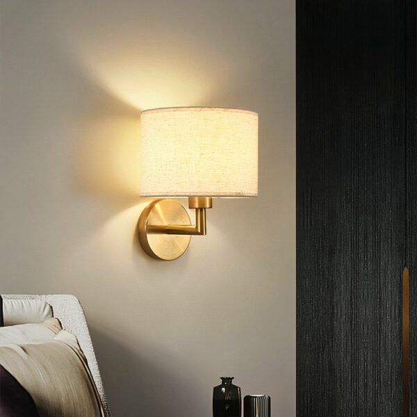 LED-Wandleuchte aus Stoff mit goldfarbener Metallhalterung, Licht an, neben einem Bett.