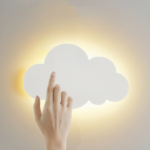 Auf einem weißen Hintergrund sehen Sie eine Wandleuchte in Form einer weißen Wolke, die eingeschaltet ist, und eine Frauenhand auf der Wolke.