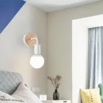 Weiße Wandleuchte mit Holzsockel und sichtbarer Glühbirne über einem Bett