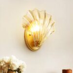 Auf einer weißen Wand sehen Sie eine Wandapplikation in Form einer Muschel, die goldfarben und transparent ist. In der linken unteren Ecke sehen Sie einen Strauß weißer Rosen.
