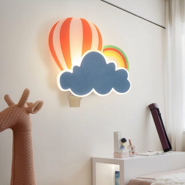 An der Wand eines Kinderzimmers sehen Sie in der Mitte des Bildes eine Wandleuchte in Form einer Wolke, eines Heißluftballons und eines Regenbogens für das Kinderzimmer. In der unteren linken Ecke des Bildes befindet sich ein rosafarbener Giraffenspielzeugkopf.
