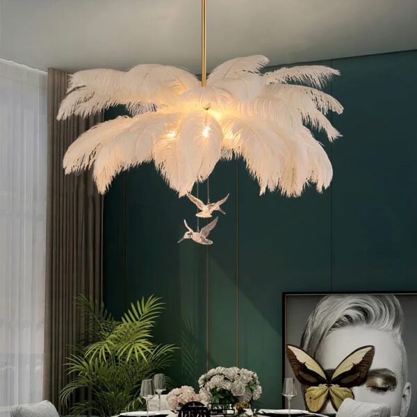 Kronleuchter avec grandes plumes blanches, et deux figurines transparentes d'oiseaux. Suspendus. Les ampoules sont allumées et camouflées dans les plumes. On voit un mur vert foncé dans le fond, ainsi qu'un tableau et une plante verte.