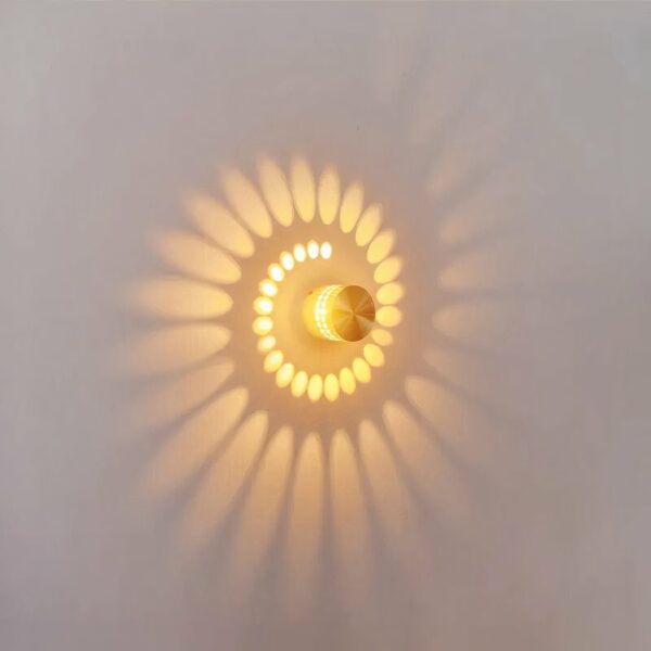 Applique murale a effet, se compose d'un cylindre métalique gris, perferé de petit trous, qui laisse diffusé la lumière en forme de spirale au mur. Allumée. Accroché sur un mur blanc.