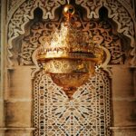 Grand lustre doré marocain, majestueux, en haut plusieurs couronnes qui donnent l'effet des flemmes, le bas descend en pointe. Tout le lustre est évidé et ajouré. Derrière un mur typique marocain avec une partie en bois sculpté et l'autre des pierres et des carreaux aux motifs marocains.