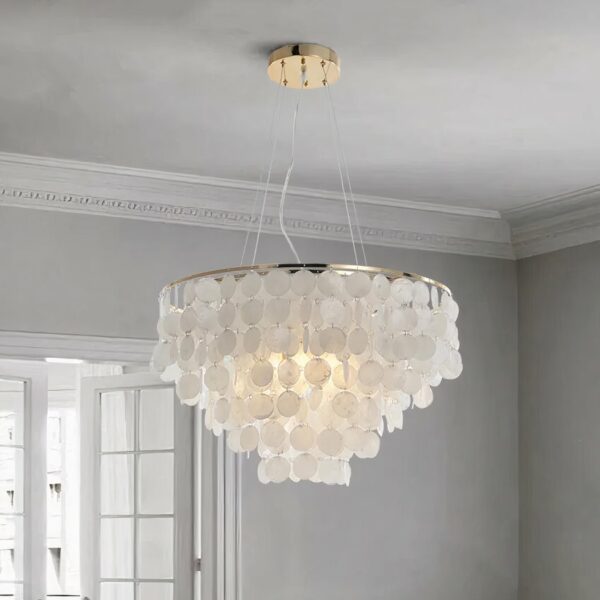 Suspension ronde avec pleins de perles de nacre ronde et plates suspendues en cascade en forme conique. On voit un plafond gris clair, et le mur du fond gris clair avec une porte-fenêtre blanche à carreaux.