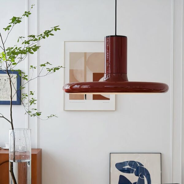 Kronleuchter rouge rond, plat vintage, suspendu. Au fond mur blanc avec deux tableaux et à gauche un vase haut avec de l'eau et une plante verte.