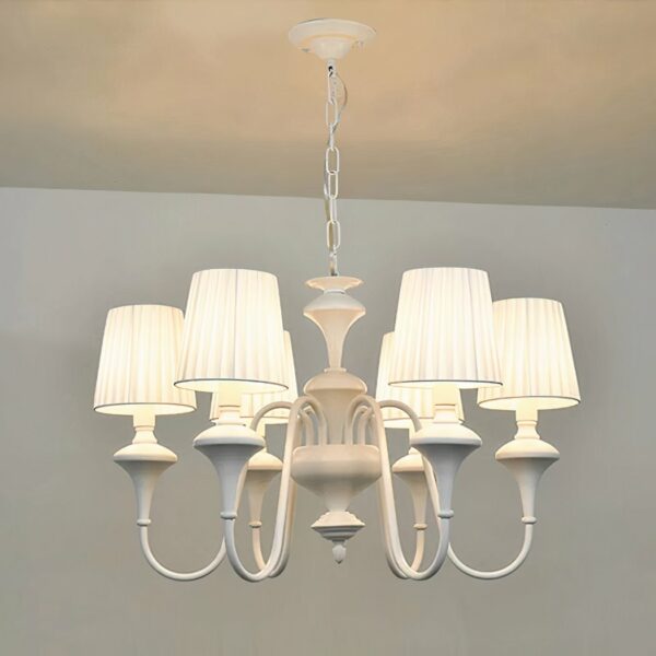 Kronleuchter blanc style baroque, en forme de chandelier avec 6 abat-jours blanc, les ampoules sont allumées. Suspendu par une chaine à un plafond blanc, on voit un bout de mur blanc dans le fond.
