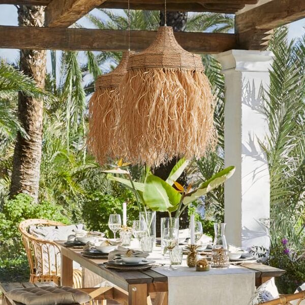 Handgeflochtener Kronleuchter aus handgefertigtem Stroh, der unter einer Pergola und über einem Esstisch inmitten üppiger Bäume hängt.