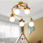 Kronleuchter für ein Kinderzimmer, in Form einer braunen Spirale, an der 5 Tierköpfe hängen und darunter die eingeschaltete Glühbirne: Giraffe, Zebra, Träumer, Tiger, Kuh. Links ist ein Fenster mit weißem Schleier, rechts eine beigefarbene Wand. Dazwischen das Dach eines Kinderhauses aus Stoff.