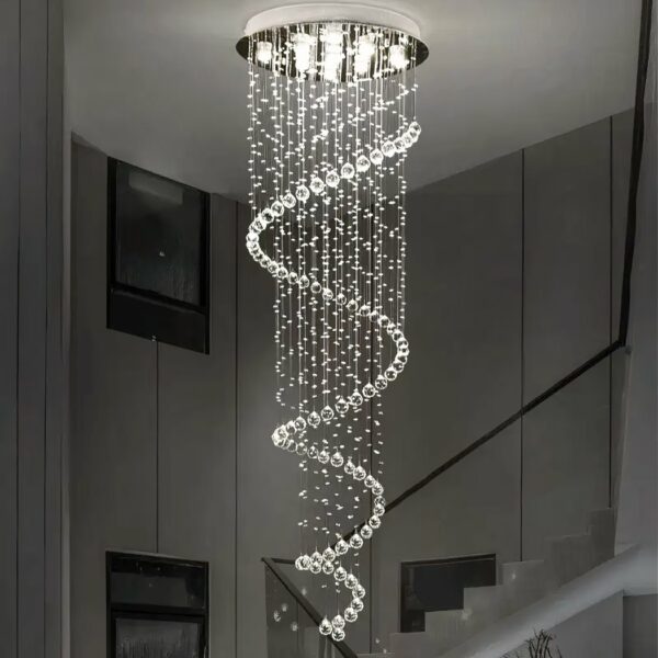 Deckenkronleuchter in Form einer Spirale aus wassertropfenförmigen Aufhängungen. Weißes Licht wird von mehreren LEDs in der runden, silbernen Halterung an der Decke eingeschaltet. Dahinter befindet sich eine Treppe mit schwarzem Geländer und Bildern an der Wand.