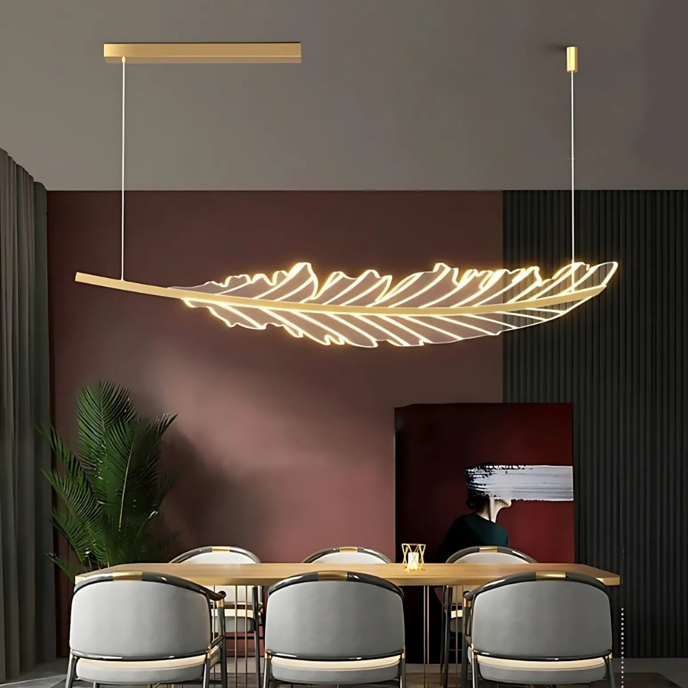 Lustre en forme de feuille suspendue en acrylique avec un support fixé au plafond de couleur doré. Il est au-dessus d'une table rectangulaire en bois avec 6 chaises grises. Au fond un mur noir et bordeaux avec un tableau peint. Sur la gauche, une plante style palmier et un rideau gris.